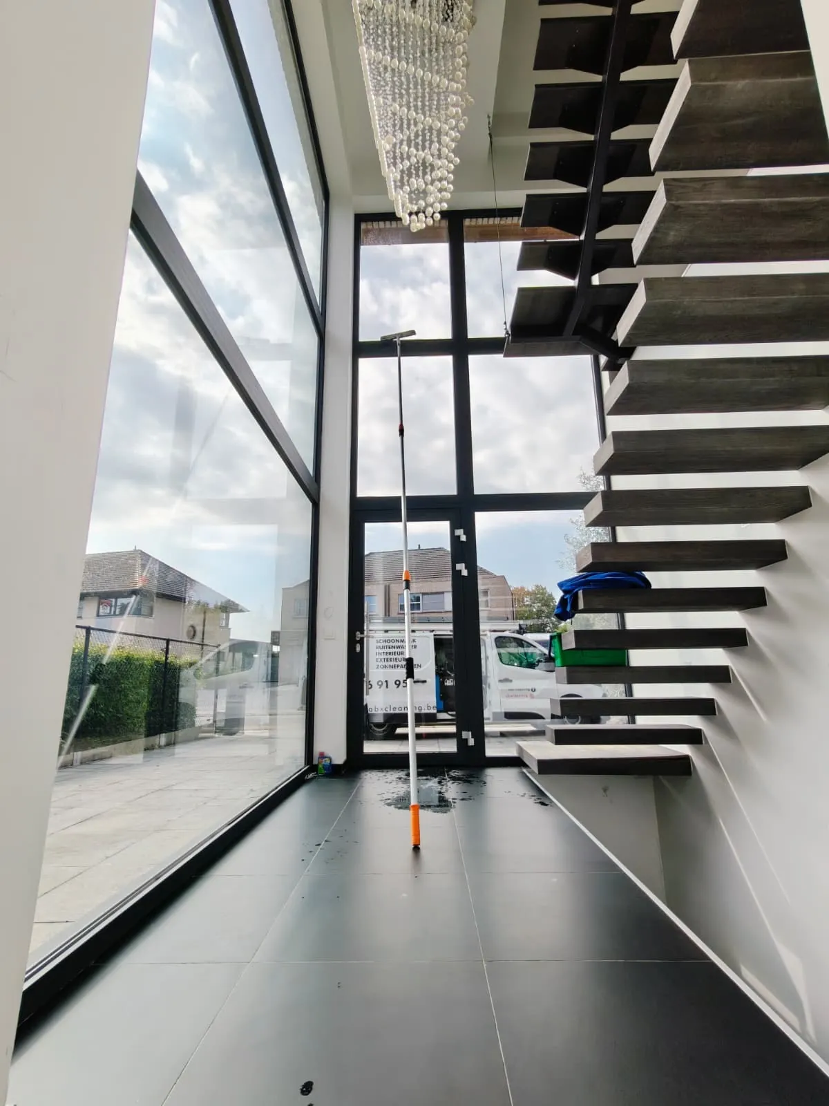 Hoge glaswand in een woning, aan de binnenkant gereinigd met een telescopische steel en trekker. Buiten staat de bestelwagen van ABX Cleaning.